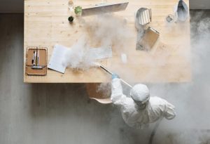 Person in protective suit steaming a desk with office supplies
